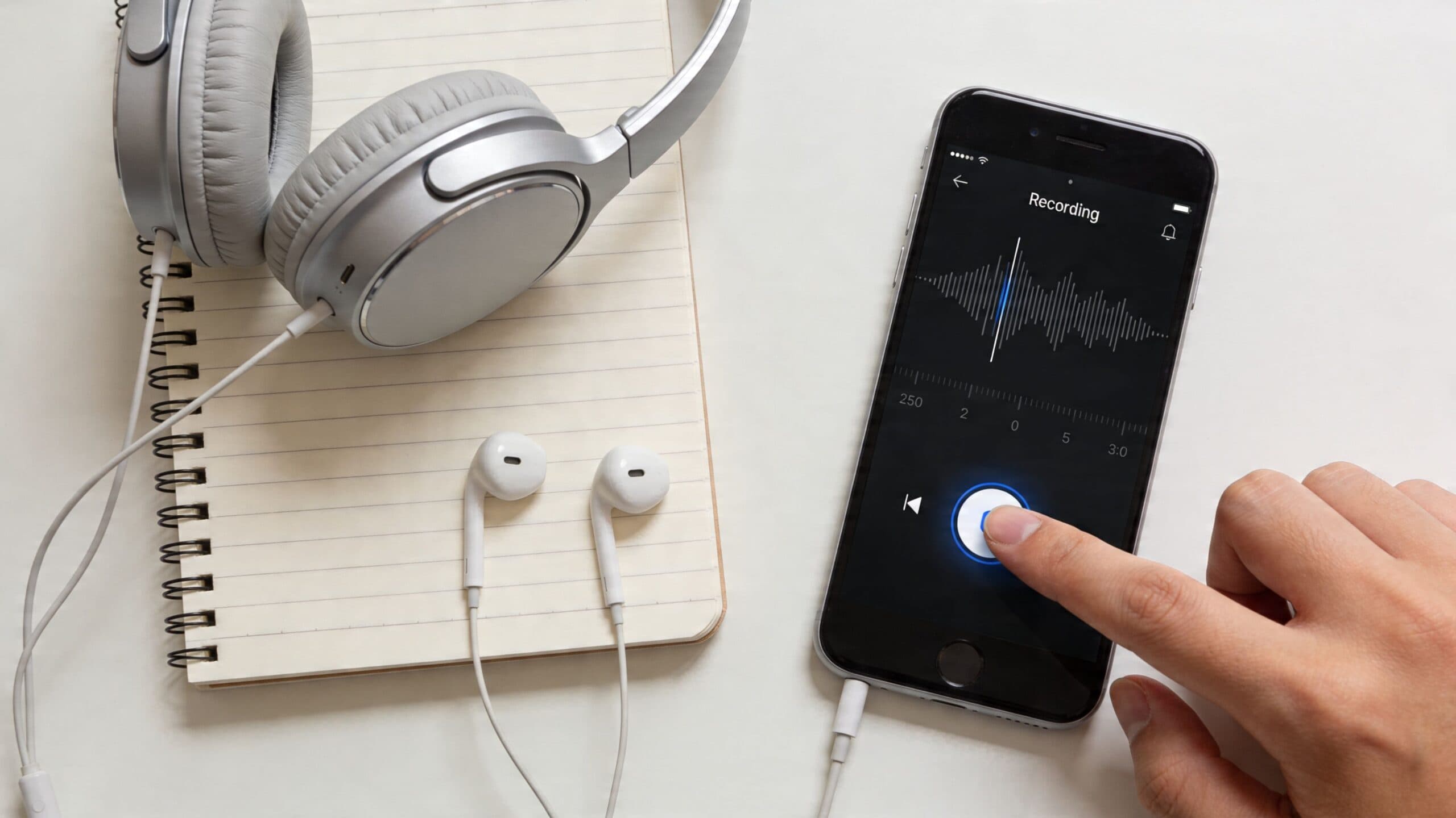 A hand pressing the record button on a smartphone with headphones and a notebook on a desk.